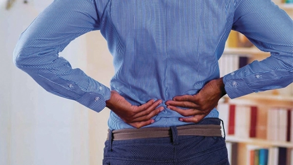 Person standing indoors with both hands placed on the lower back, highlighting the lumbar area.